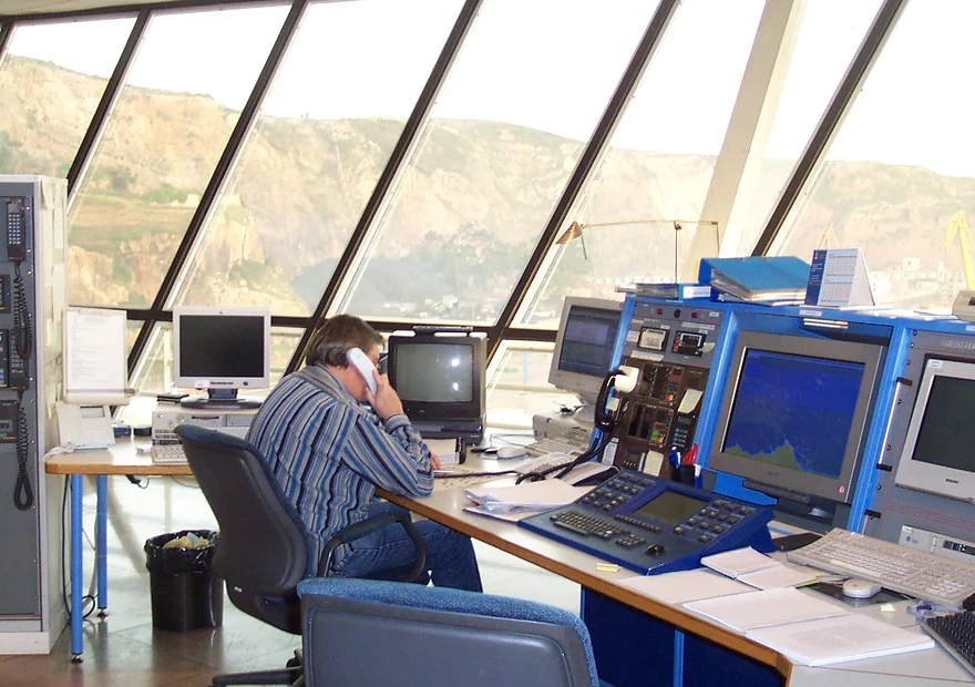 Operador de un centro de control de tráfico marítimo hablando por teléfono ante las consolas de radar, con el ventanal sobre la costa al fondo