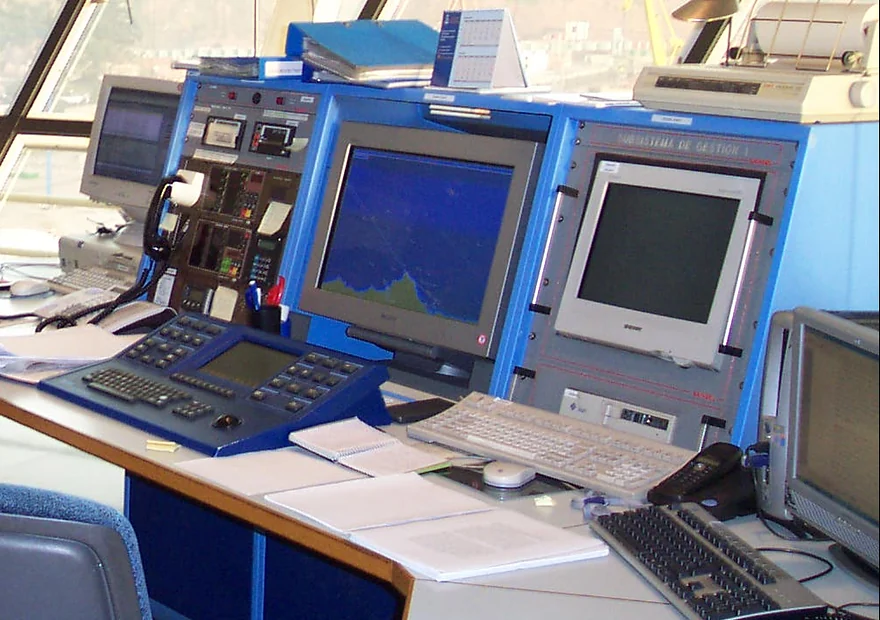 Consolas y pantallas de radar de un puesto de operador de tráfico marítimo, con los teclados y el cuaderno de servicio sobre la mesa